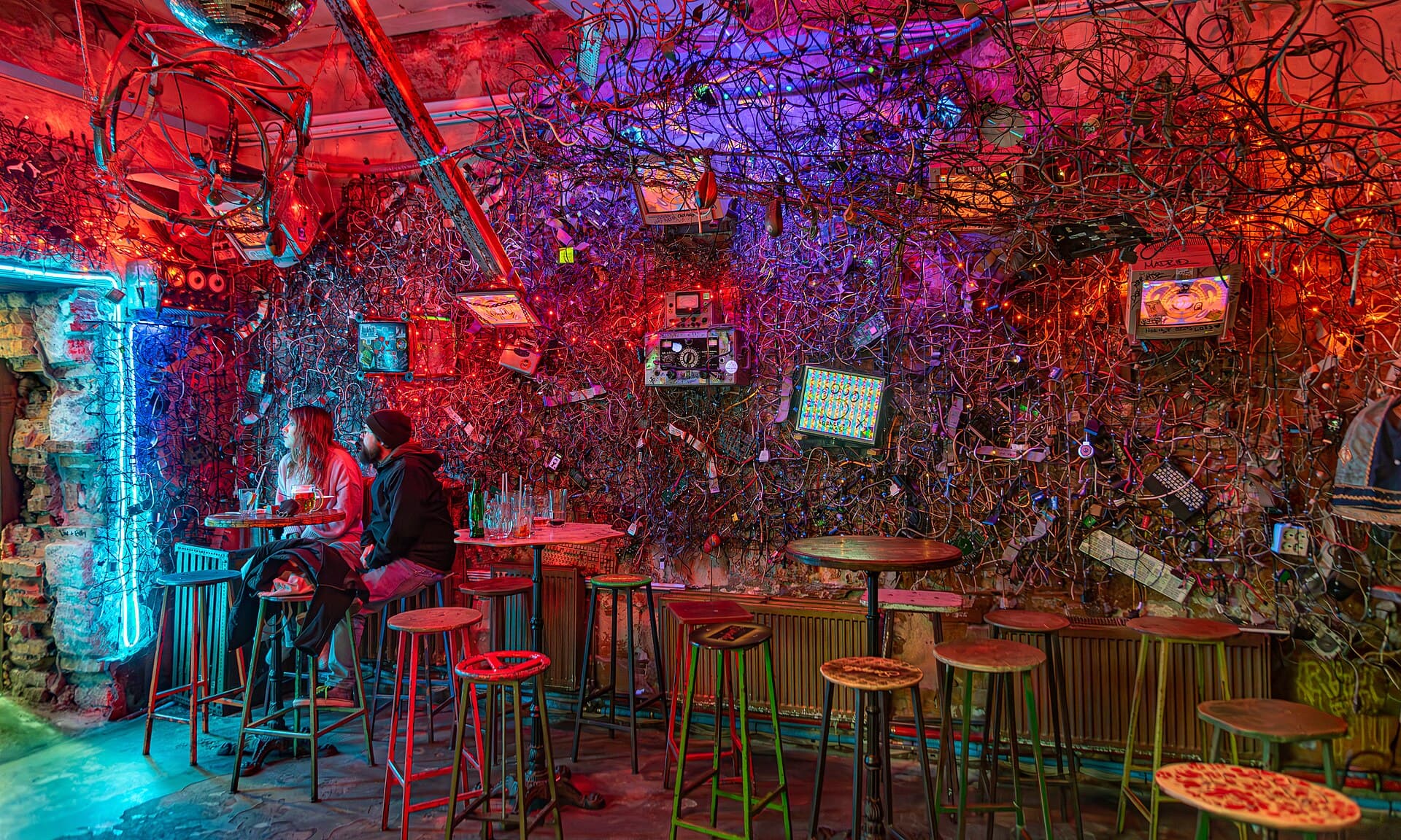 An interior room at Szimpla Kert lit in red and purple neon, its walls and ceiling completely covered in tangled cables, old monitors, keyboards and dismantled electronics, with two people sitting at a table with beers
