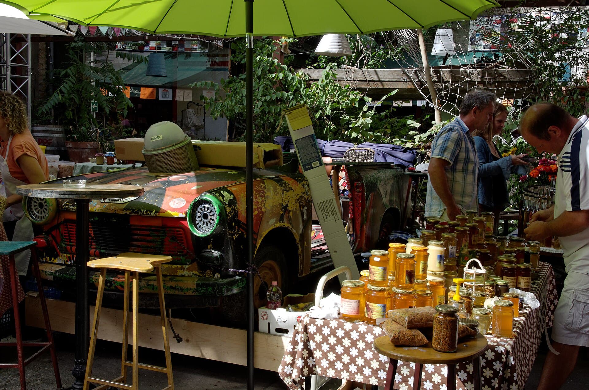 The Szimpla Kert farmers market in daylight, with a stall of honey jars in the foreground under a green parasol, a beekeeper's hat resting on the bonnet of the painted Trabant, and producers serving shoppers