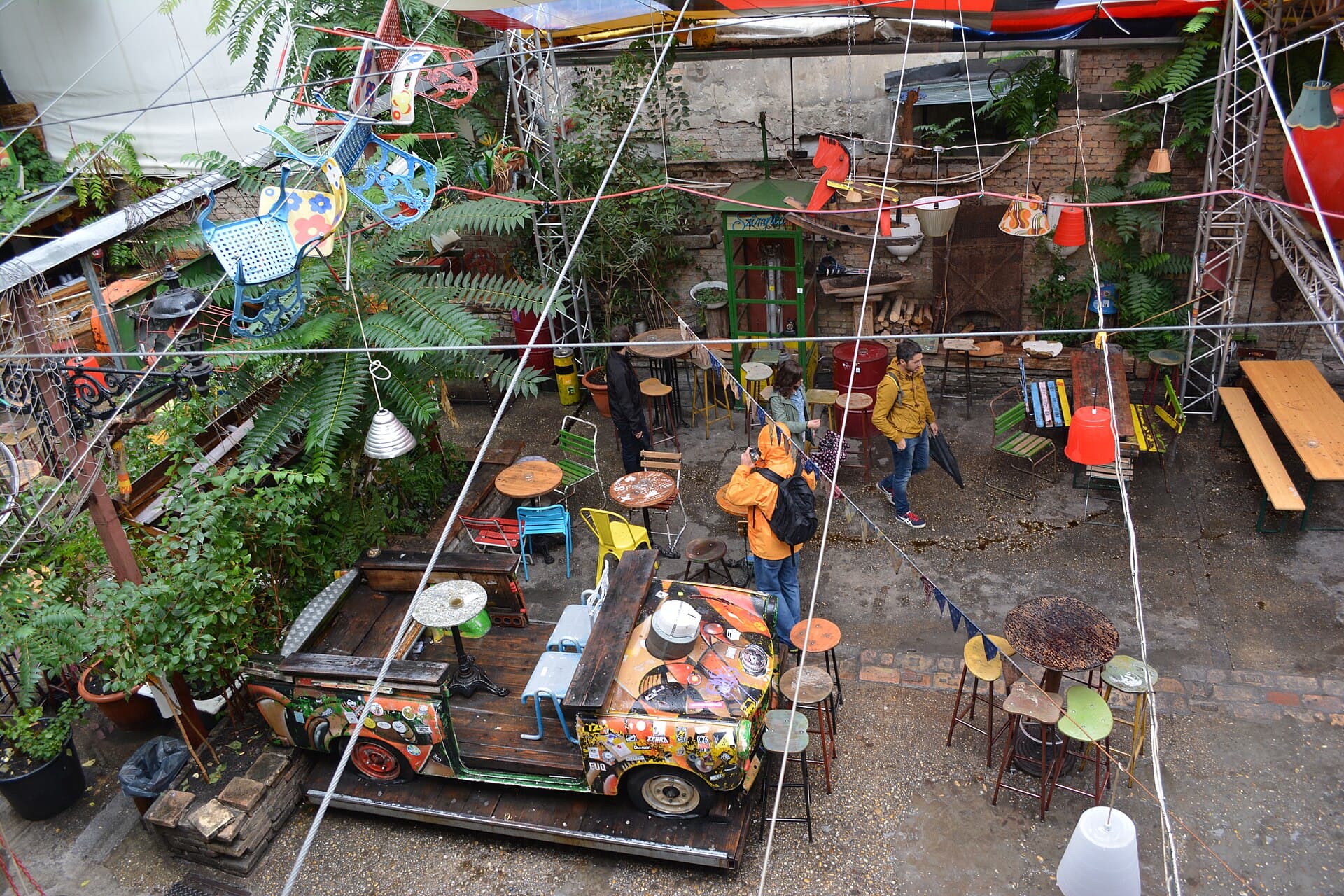 The Szimpla Kert courtyard in daylight and nearly empty, seen from the gallery above, with the sticker-covered Trabant car on a wooden platform, painted chairs strung overhead on wires and a scattering of mismatched stools