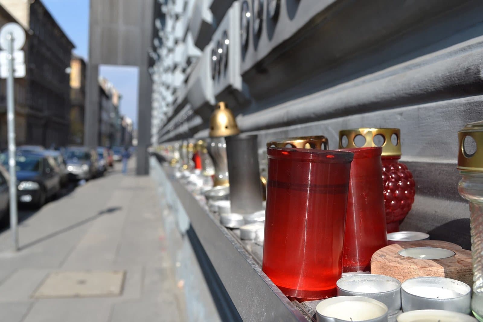 Memorial candles in red and clear glass jars with gold lids, lined up along the granite ledge at the base of the House of Terror, with the black steel wall of the building rising above them and Andrassy Avenue receding into the distance behind