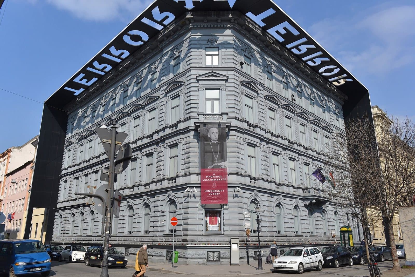 The House of Terror on the corner of Andrassy Avenue in Budapest: a grey neo-Renaissance apartment block framed by a black steel canopy that projects over the roofline, with the word TERROR cut out of the overhang in large letters, a banner hanging on the corner facade, and parked cars and pedestrians on the street below