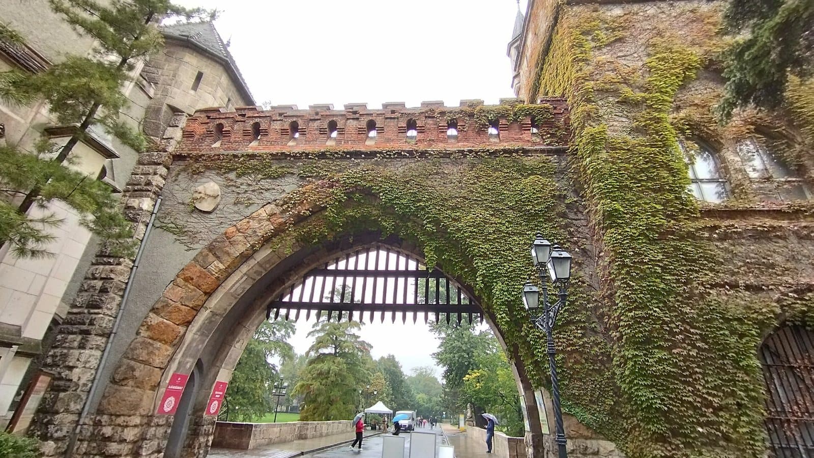 The gatehouse of Vajdahunyad Castle in Budapest seen from below in the rain: a stone arch with a raised wooden portcullis, a crenellated brick wall walk above it, dense green ivy covering the tower to the right, an ornate iron street lamp, and people with umbrellas walking through towards the park